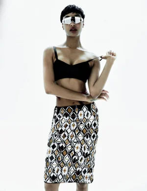 Jourdan Dunn wearing futuristic wraparound sunglasses, black bralette, and patterned skirt against white background