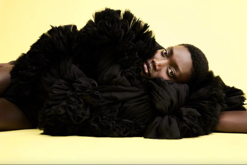 Model Fatou Kebbeh wearing a voluminous black ruffled dress lying against a pale yellow background.