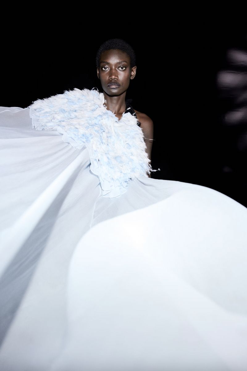 Runway fashion shot of a Black female model with short hair wearing a white flowing feathered dress against a dark background.