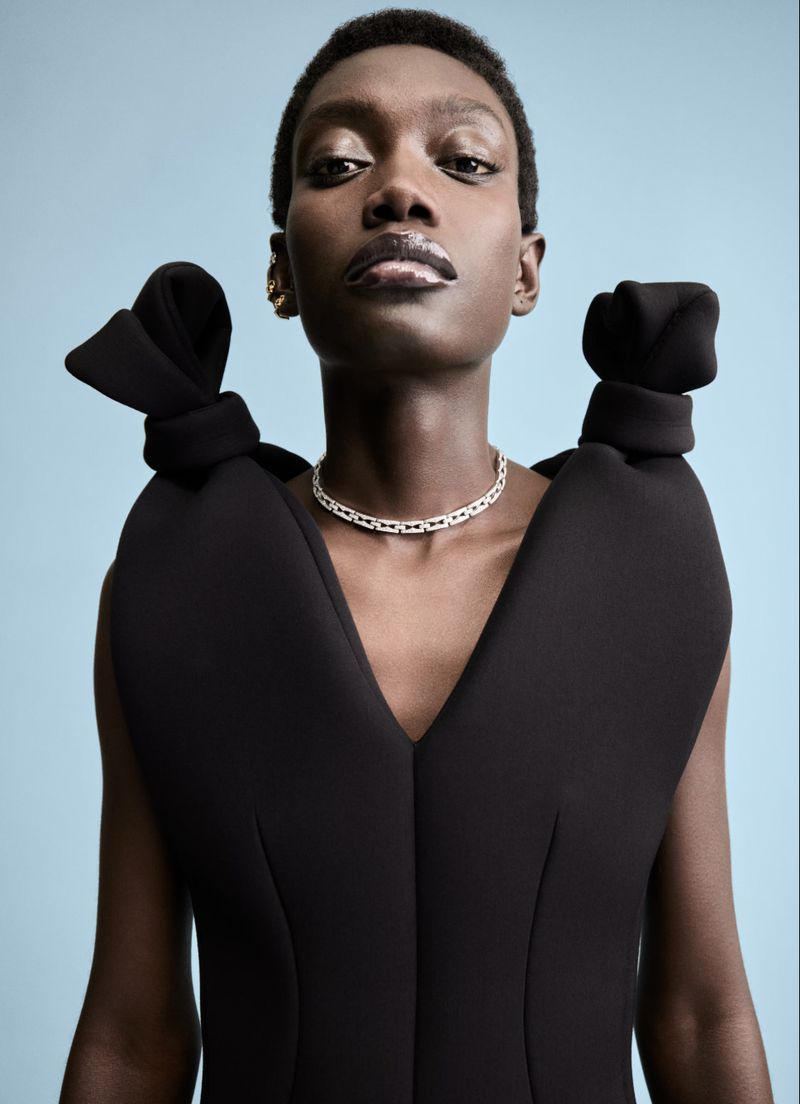 Model Fatou Kebbeh wearing a black dress with knotted shoulder details against a blue backdrop