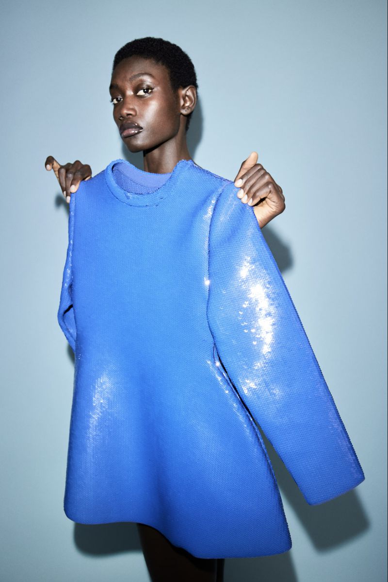 An adult Black female model poses in a studio with a bright blue, sequined, long-sleeved dress held behind her by another person.