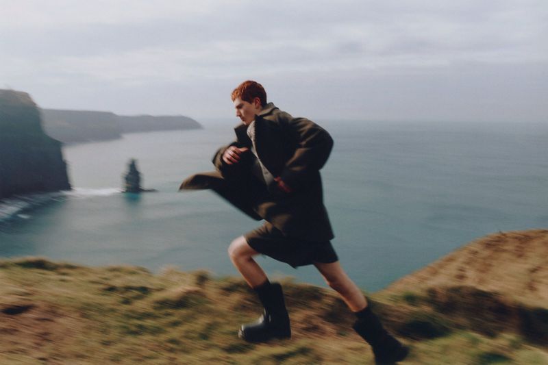 Man in coat runs on grassy cliff overlooking ocean with rock formations. Motion blur creates a sense of speed against the cloudy sky.