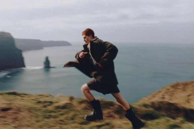 Man in coat runs on grassy cliff overlooking ocean with rock formations. Motion blur creates a sense of speed against the cloudy sky.