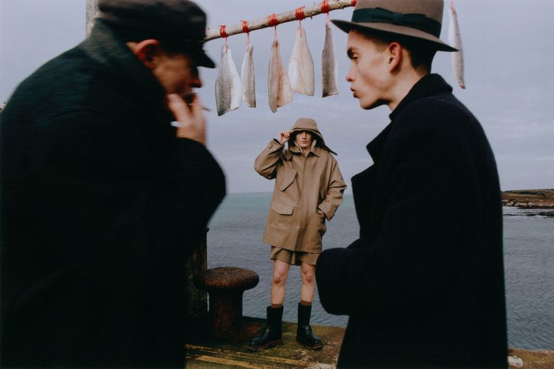 Three figures in outerwear standing on a pier, with dried fish hanging above. Coastal scene with water and overcast sky visible.