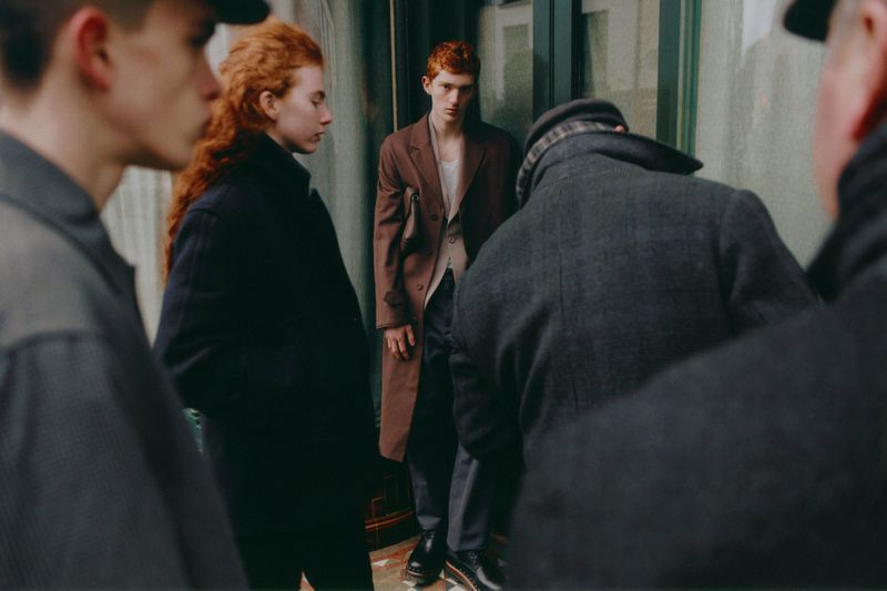 Outdoor fashion shot showcasing a redhead model in a brown overcoat and grey trousers, surrounded by blurred people in a neutral palette.