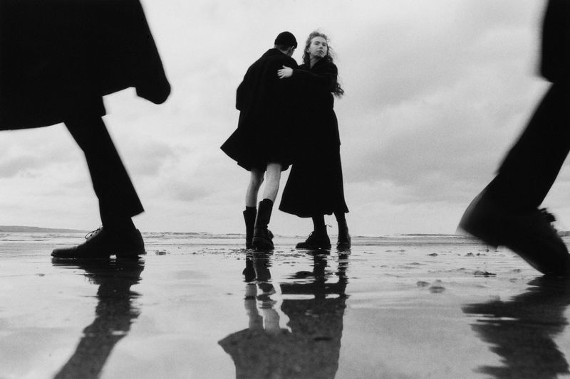 Black and white editorial fashion image featuring two figures embracing on a wet sandy beach under an overcast sky, framed by blurred silhouettes.