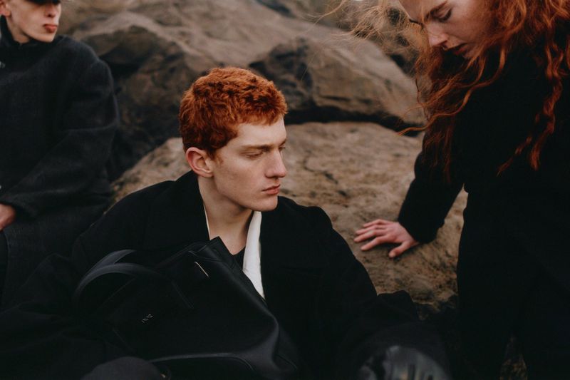 Fashion image featuring a young adult man with curly red hair wearing a black coat, posed against a rocky background with other models.