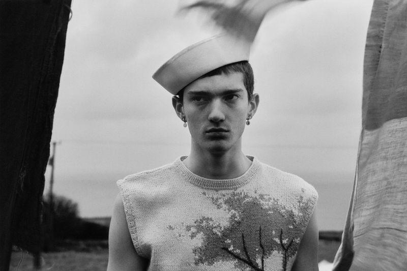 Monochrome portrait of a young adult male wearing a knitted top, hat and earrings, outdoors against an overcast sky and distant horizon.