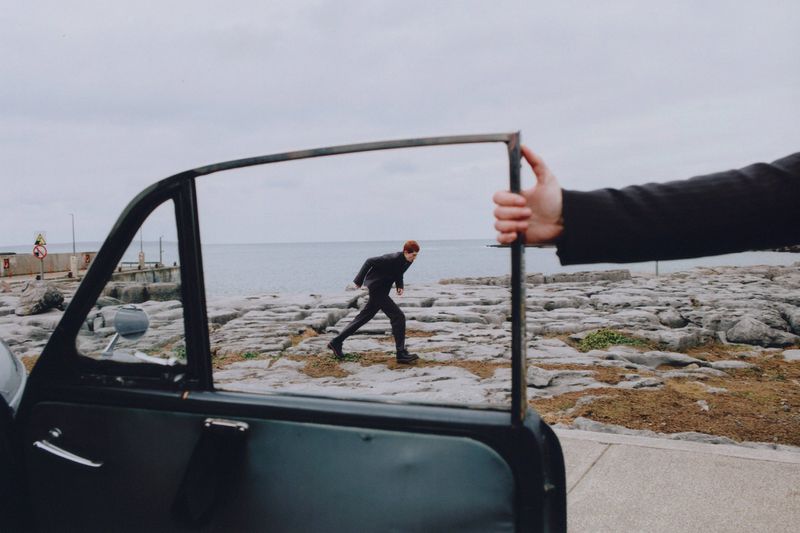 A man in dark clothing running on a rocky beach towards the ocean, viewed through the open door of a dark-colored vintage car.