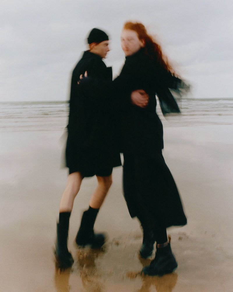 Two models pose on a wet sand beach with muted sky. One has dark hair and wears a beanie, and the other has long red hair.