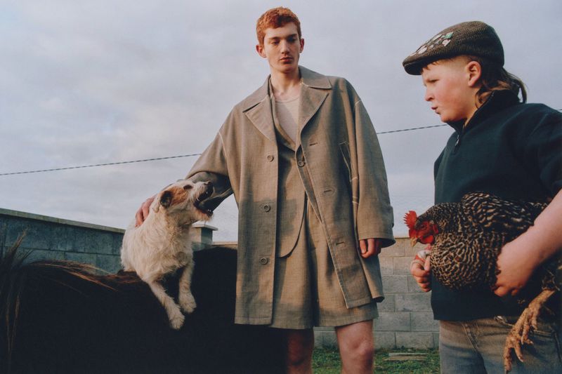 Two children pose outdoors with farm animals: a boy holds a chicken, another pets a dog on a horse, under a cloudy sky.