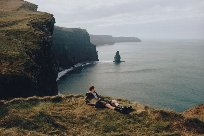 Adult lying on grassy cliffside overlooking the sea, distant cliffs and rock formations, cloudy skies, creating a peaceful travel image.