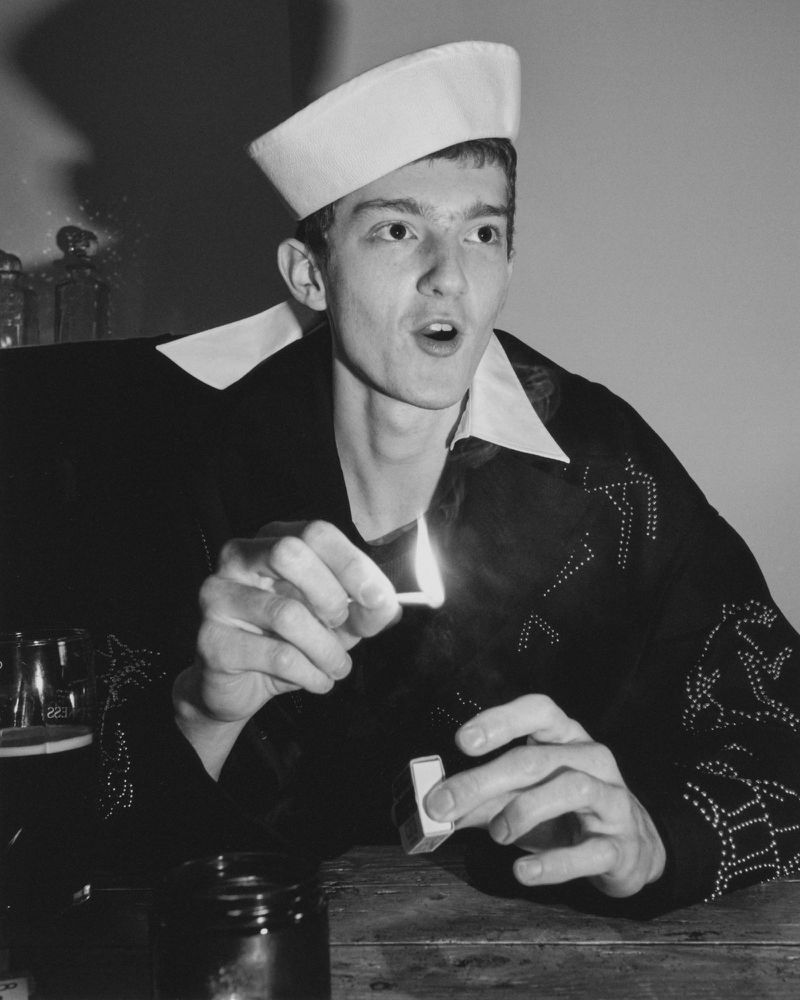 Monochrome portrait of a young adult in a sailor cap lighting a match, showcasing vintage fashion in an indoor setting.