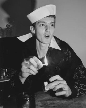 Monochrome portrait of a young adult in a sailor cap lighting a match, showcasing vintage fashion in an indoor setting.