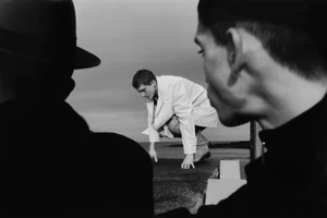 Black and white editorial fashion image featuring two men in silhouette foreground watching another man crouching outdoors near water.
