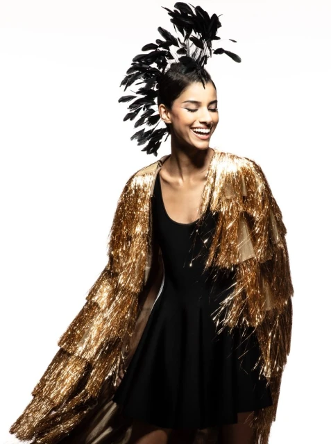Smiling woman in a black dress and gold tinsel coat with a black feather headdress posing against a bright white background.