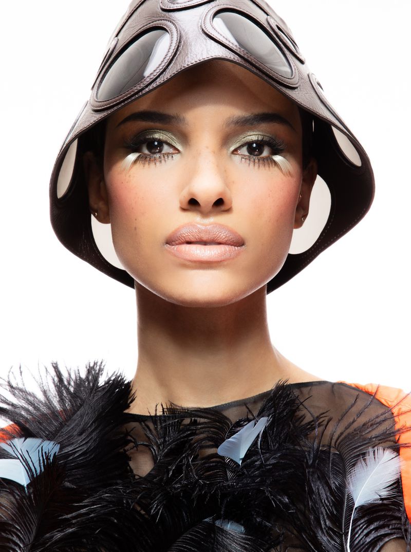 Close-up portrait of a woman wearing a unique brown leather hat with circular cutouts and a black feathery top against a white background.