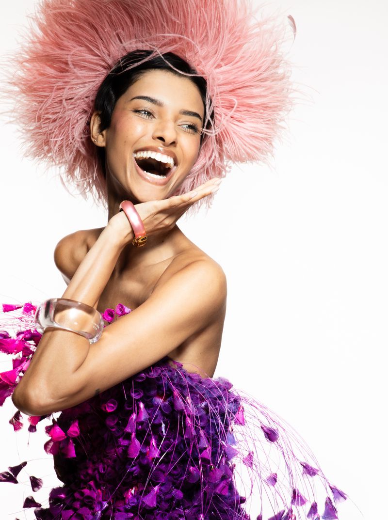 Smiling woman poses in a purple and pink feathered dress with a feathered headpiece against a white backdrop in a studio shot.