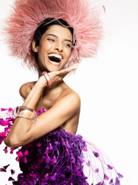 Smiling woman poses in a purple and pink feathered dress with a feathered headpiece against a white backdrop in a studio shot.