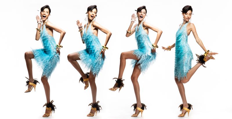 Four women dance in blue fringe dresses with feather-embellished heels on a white background, showcasing energetic fashion poses.