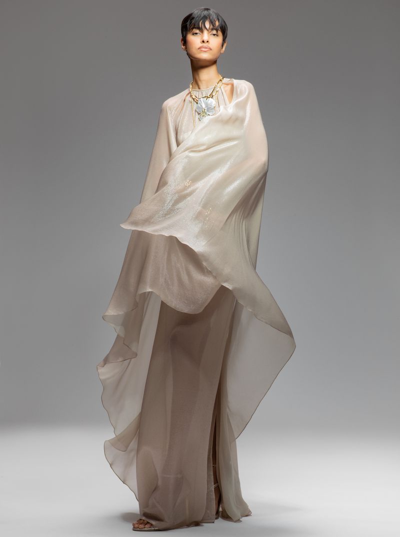 Full-length portrait of a woman in a flowing, light-colored gown and statement necklace posing in a bright, neutral studio setting.