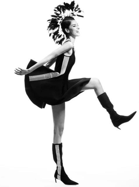 A slender woman in a black dress and boots poses dynamically with a feathered headdress against a stark white background in a fashion editorial.