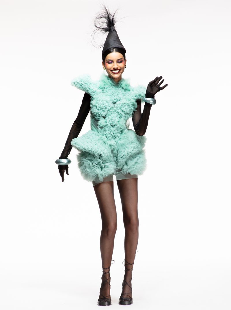 A full-length studio shot features a smiling woman in a sculptural, textured light-green dress with matching bracelets and a pointed black hat.
