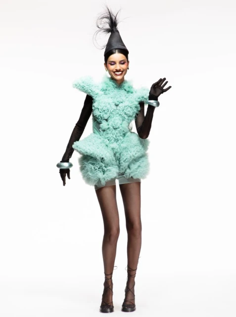 A full-length studio shot features a smiling woman in a sculptural, textured light-green dress with matching bracelets and a pointed black hat.