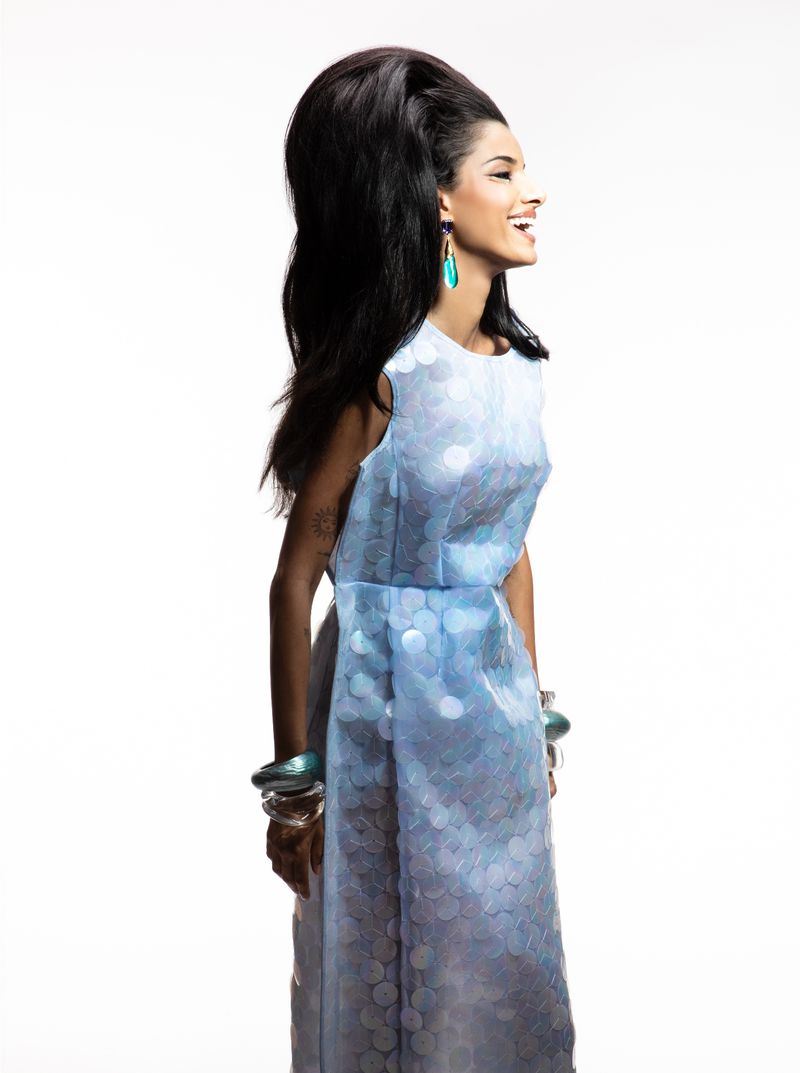 A woman with dark hair and turquoise earrings wears a light blue dress with matching bracelets against a white background in studio lighting.