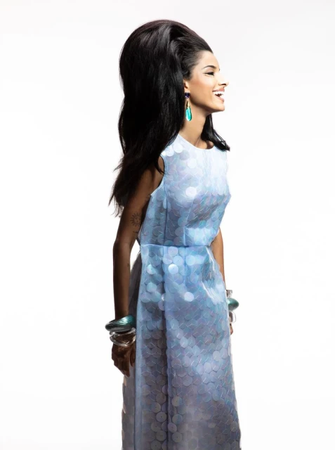 A woman with dark hair and turquoise earrings wears a light blue dress with matching bracelets against a white background in studio lighting.