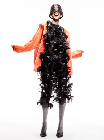 Fashionable woman wearing a unique helmet, an orange jacket, and a feather boa, standing against a bright white studio backdrop.