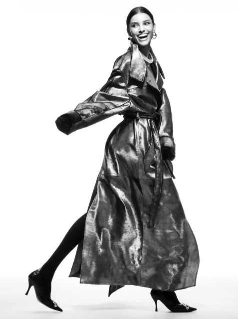 Elegant woman in a metallic trench coat strikes a pose while walking against a clean white backdrop in a studio fashion shot.
