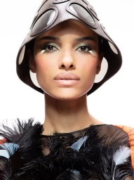 Model wearing a dark leather hat with circular lenses and a black feathered garment against a plain white background.