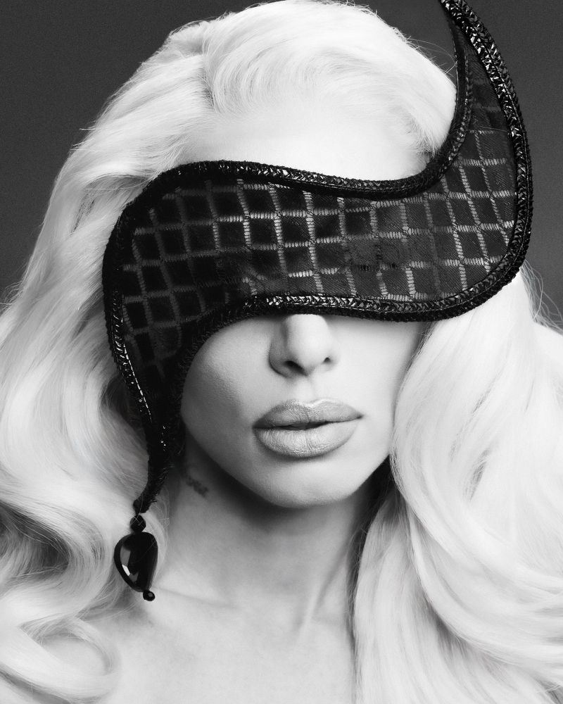 Black and white portrait of a blonde adult female wearing a black textured blindfold, looking at the camera, against a dark background.