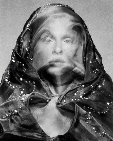Monochrome portrait of a woman with a gauzy hooded head covering and beaded embellishments, presented in a high-contrast black and white.