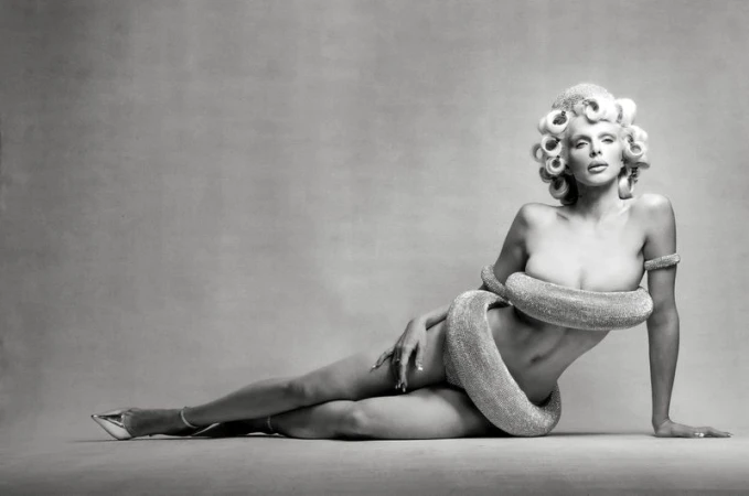 Black and white portrait of a woman with blonde curly hair in rollers and high heels posing with a snake-like object around her body.