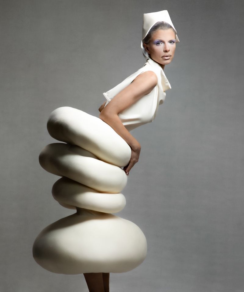 Adult female model in avant-garde fashion stands in a studio. The cream-colored sculptural dress features stacked, rounded shapes.