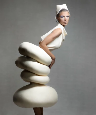 Adult female model in avant-garde fashion stands in a studio. The cream-colored sculptural dress features stacked, rounded shapes.