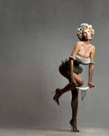 Glamour shot of a light-skinned woman in silver bikini bottom and jewelry, striking a pose in a studio setting with gray backdrop. High heels.