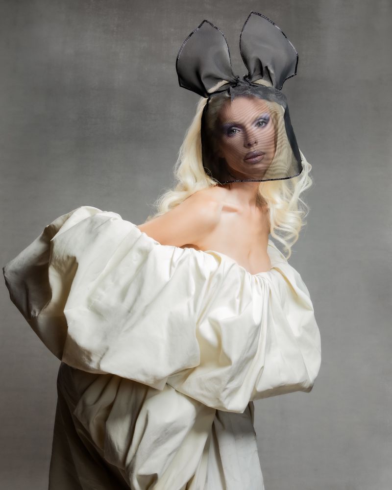 Woman in off-shoulder dress and bunny-ear headpiece poses against a grey backdrop in a fashion-forward studio portrait.