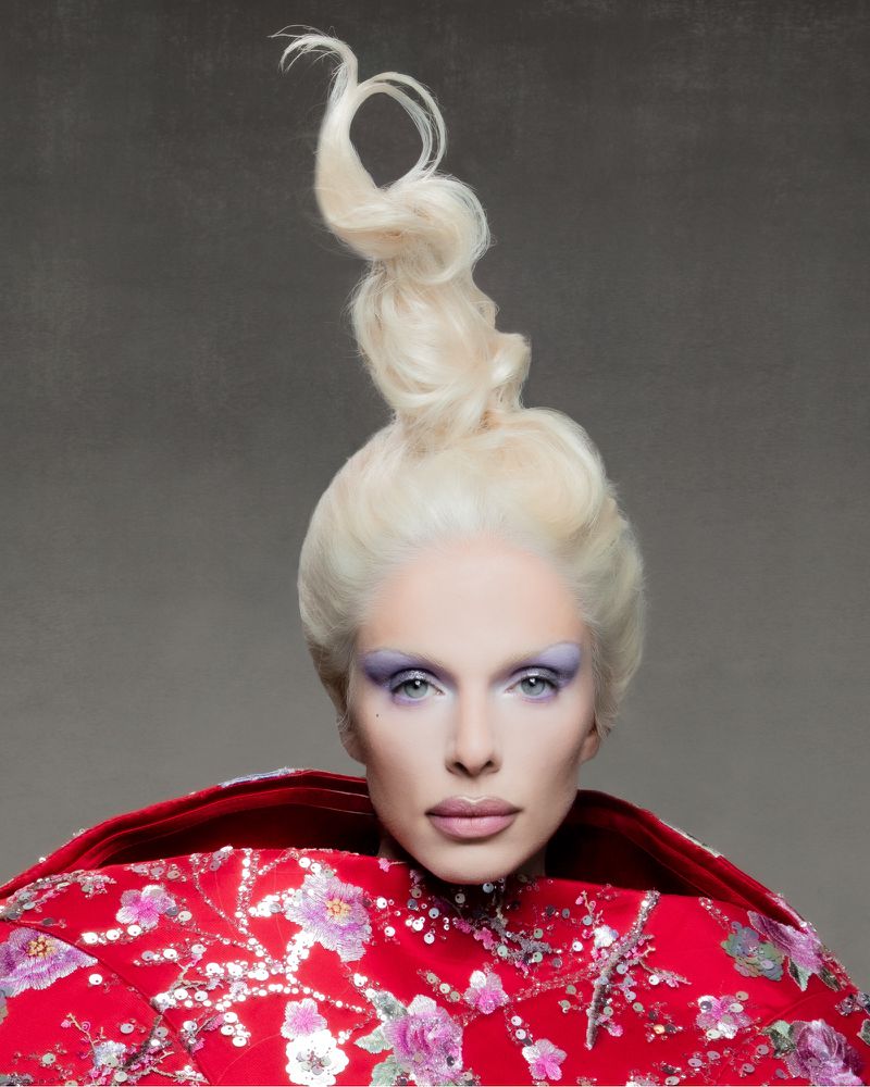 Close-up portrait of an adult woman with fair skin, wearing floral patterned red dress and dramatic white hairstyle against a grey background.