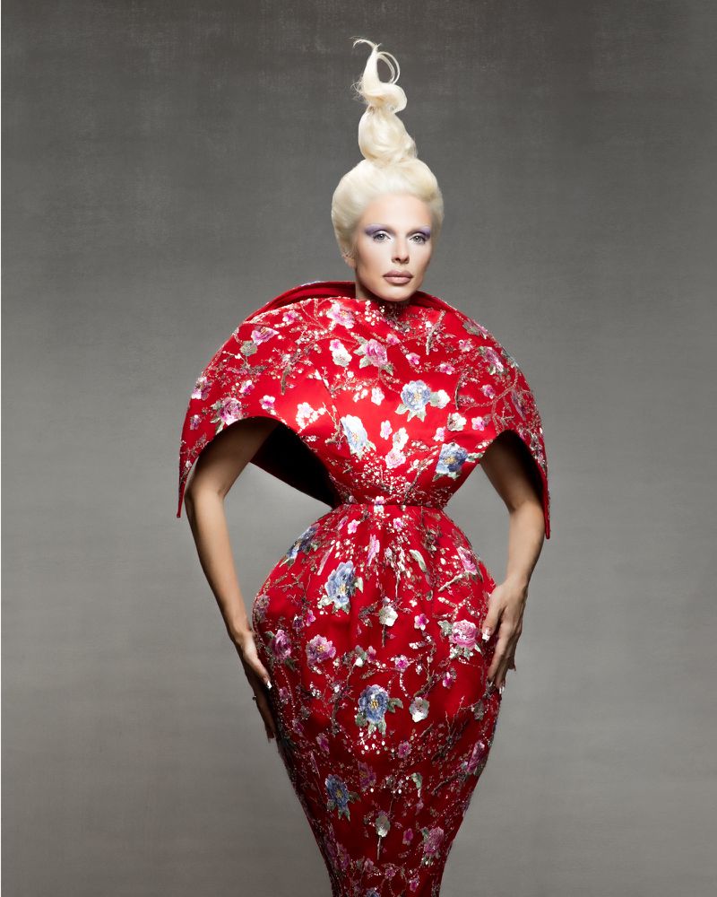 A woman with fair hair and heavy eye makeup poses in a red, embroidered gown with a wide, rounded shoulder piece on a grey background.