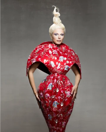 Julia Fox in a red floral structured avant garde dress with a high sculpted blonde updo against a grey background