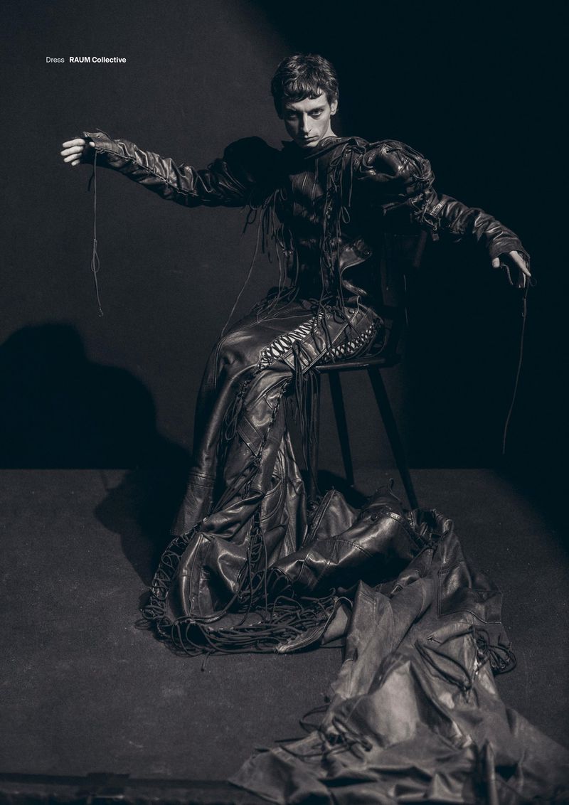 Fashion editorial shot of a young adult male wearing a dark, draped garment, seated on a stool in a studio with dramatic lighting.