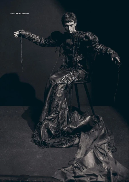 Fashion editorial shot of a young adult male wearing a dark, draped garment, seated on a stool in a studio with dramatic lighting.