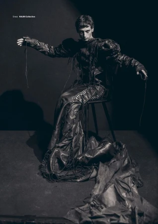 Fashion editorial shot of a young adult male wearing a dark, draped garment, seated on a stool in a studio with dramatic lighting.