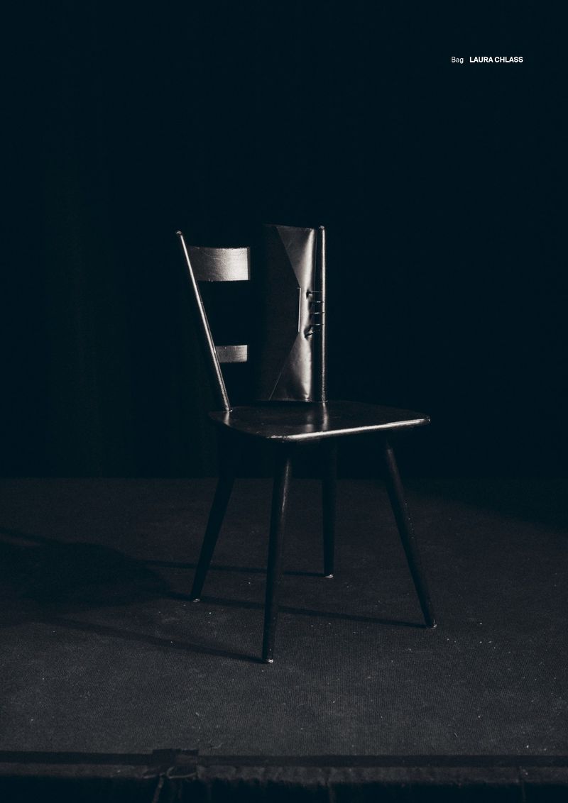 Elegant black clutch purse on a dark wooden chair against a muted black background, minimalist product-focused studio shot.