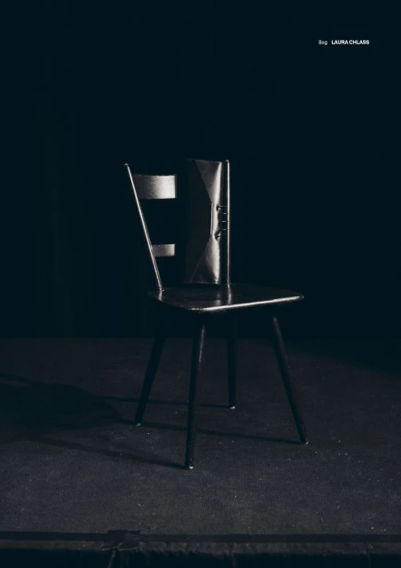 Elegant black clutch purse on a dark wooden chair against a muted black background, minimalist product-focused studio shot.