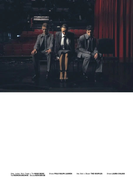 Three fashion models in suits pose seated on a stage with red curtains in the background, promoting HUGO BOSS, POLO RALPH LAUREN, THE KOOPLES, and LAURA CHIASE.