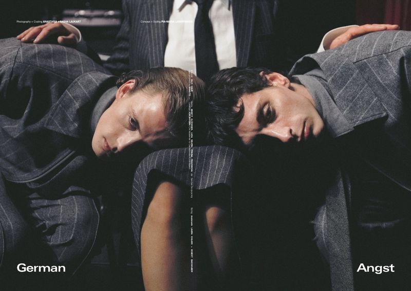 Two adult males in pinstripe suits rest their heads together. The composition creates a symmetrical, mirrored effect, and the photograph has a subdued aesthetic.